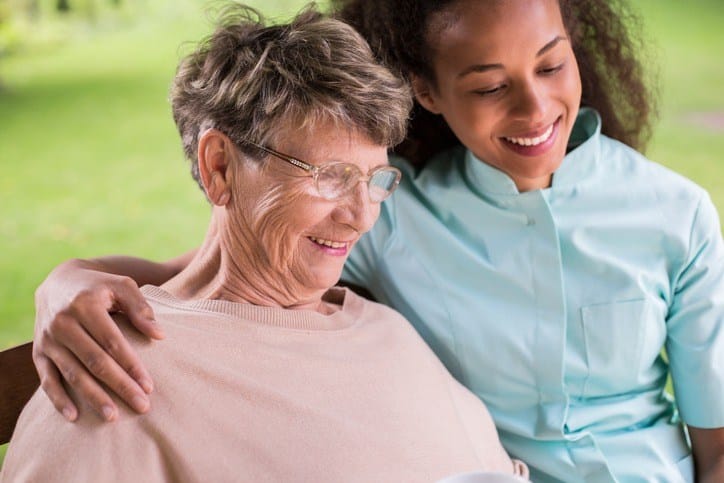 A woman sits on a bench with an older woman while in dementia care at a skilled nursing facility